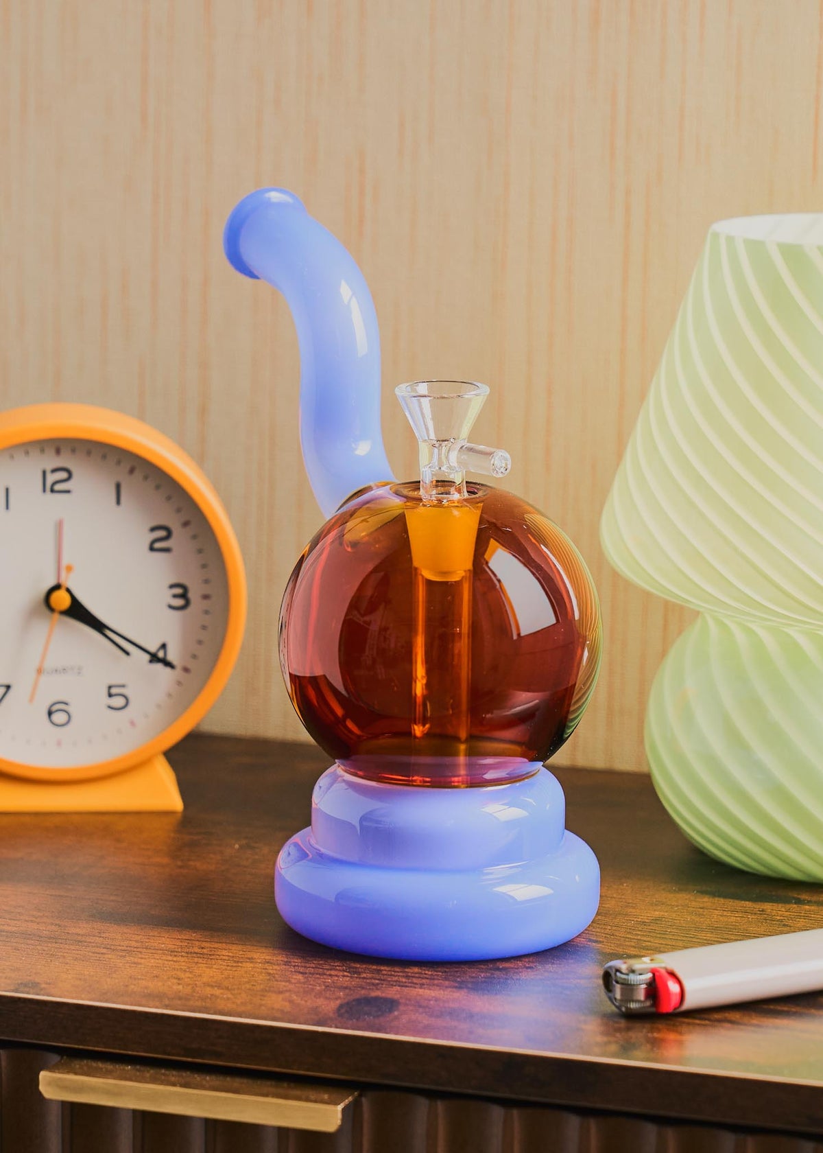 A retro-style squiggle globe bong with a bulbous amber glass body, a light jade purple curved neck and base, displayed on a wooden table next to a clock, lighter, and lamp.