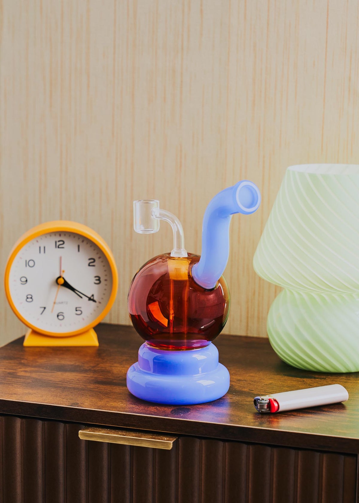 A retro-style squiggle globe rig with a bulbous amber glass body, a light jade purple curved neck and base, displayed on a wooden table next to a clock, lighter, and lamp.