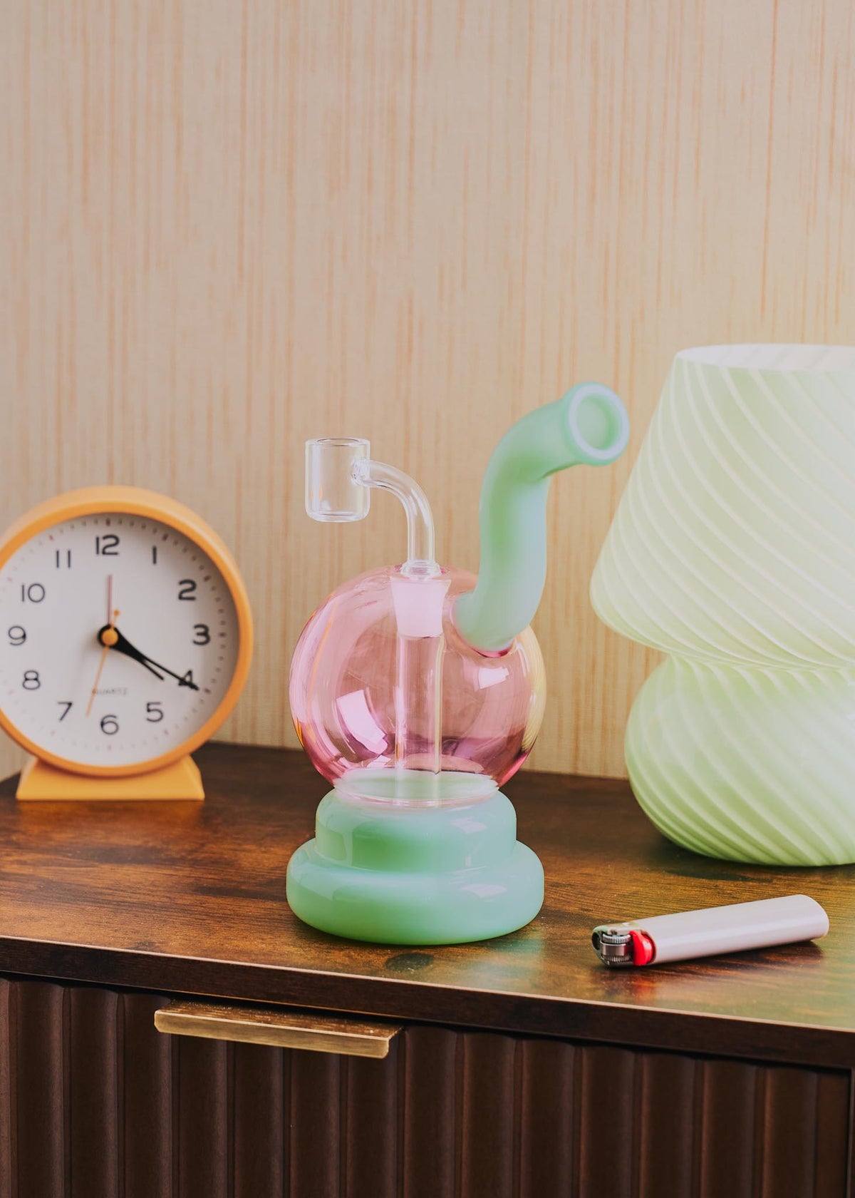 A retro-style squiggle globe rig with a bulbous pink glass body, a light jade green curved neck and base, displayed on a wooden table next to a clock, lighter, and lamp.