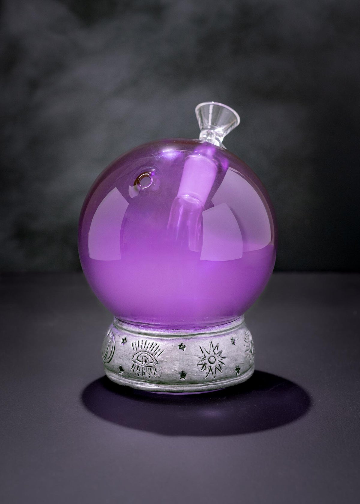 A purple crystal ball-shaped bong with a clear stopper, resting on a silver decorative base against a dark smoky background.