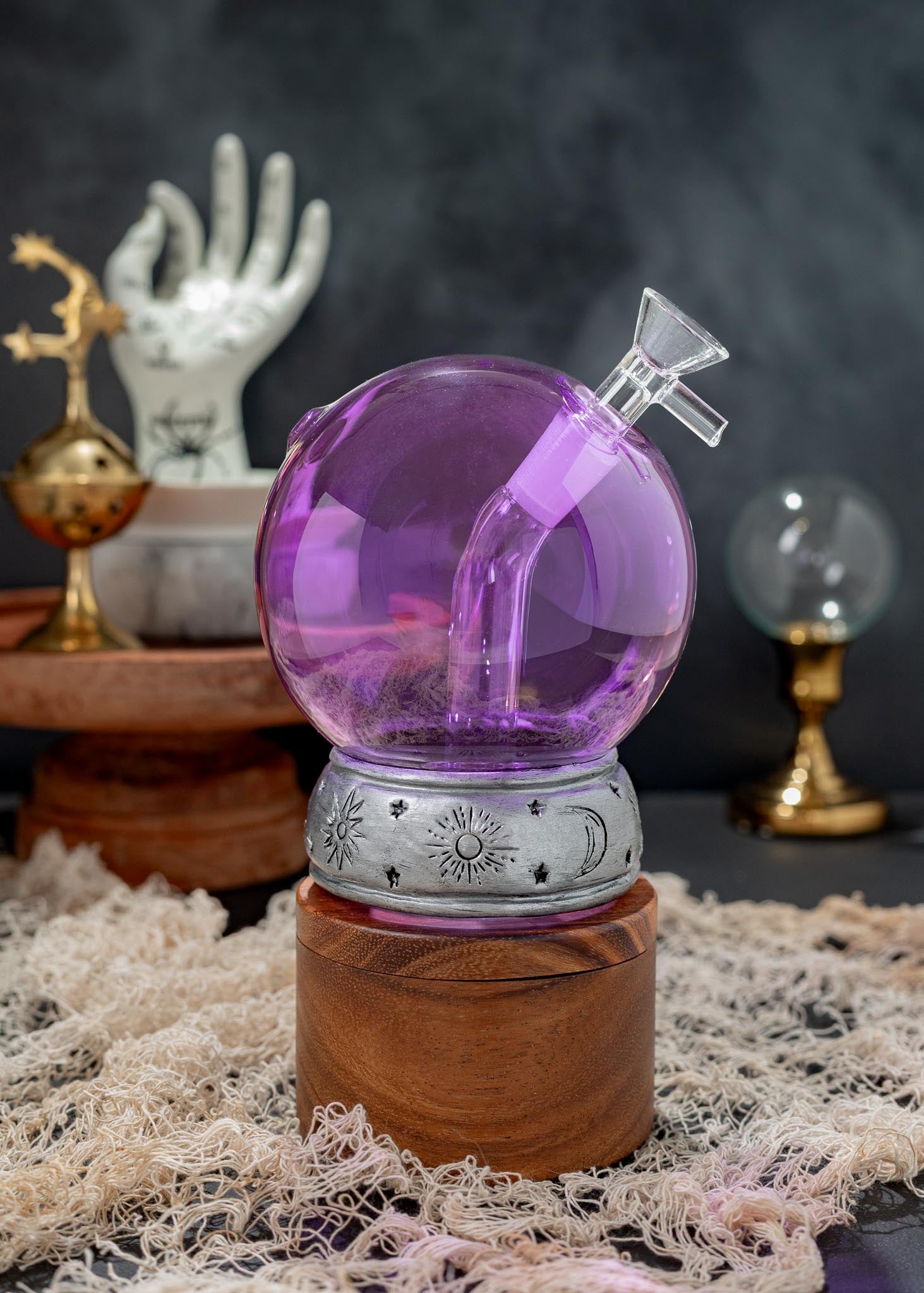 Purple crystal ball bong with a curved glass stem and silver celestial-patterned band, displayed on a wooden base with mystical props in the background.