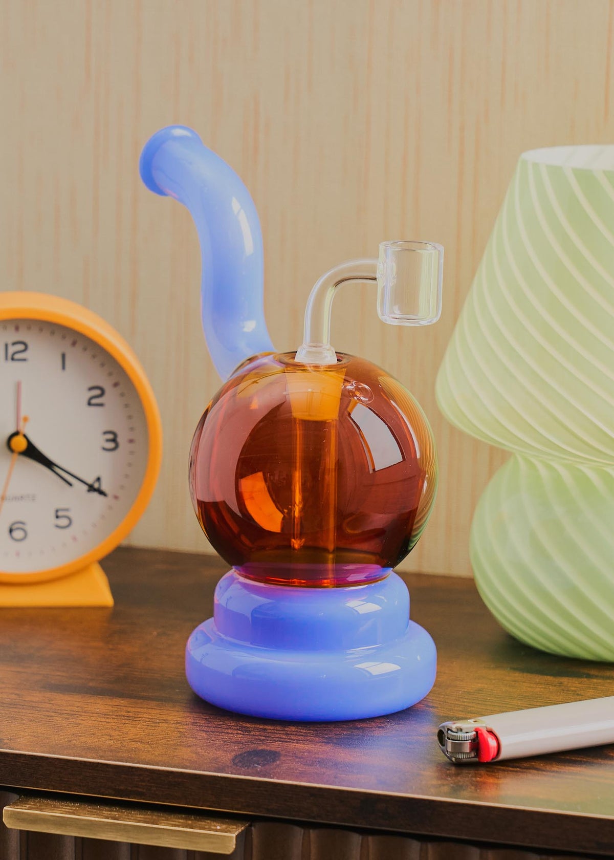A retro-style squiggle globe rig with a bulbous amber glass body, a light jade purple curved neck and base, displayed on a wooden table next to a clock, lighter, and lamp.