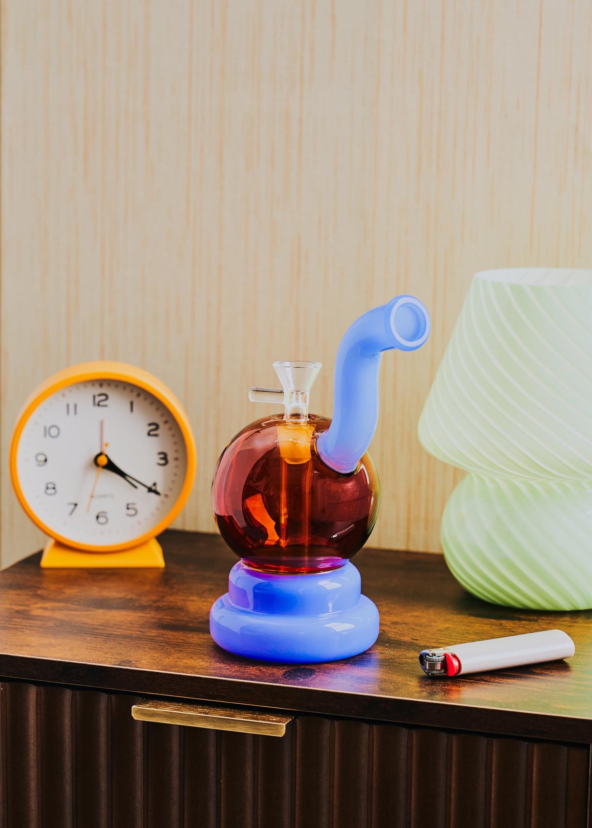 A retro-style squiggle globe bong with a bulbous amber glass body, a light jade purple curved neck and base, displayed on a wooden table next to a clock, lighter, and lamp.