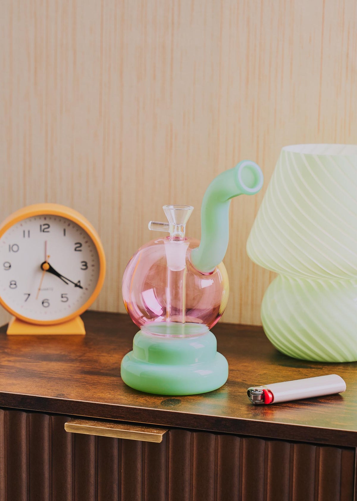 A retro-style squiggle globe bong with a bulbous pink glass body, a light jade green curved neck and base, displayed on a wooden table next to a clock, lighter, and lamp.