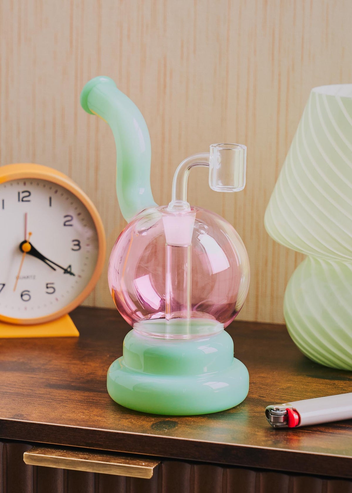 A retro-style squiggle globe rig with a bulbous pink glass body, a light jade green curved neck and base, displayed on a wooden table next to a clock, lighter, and lamp.
