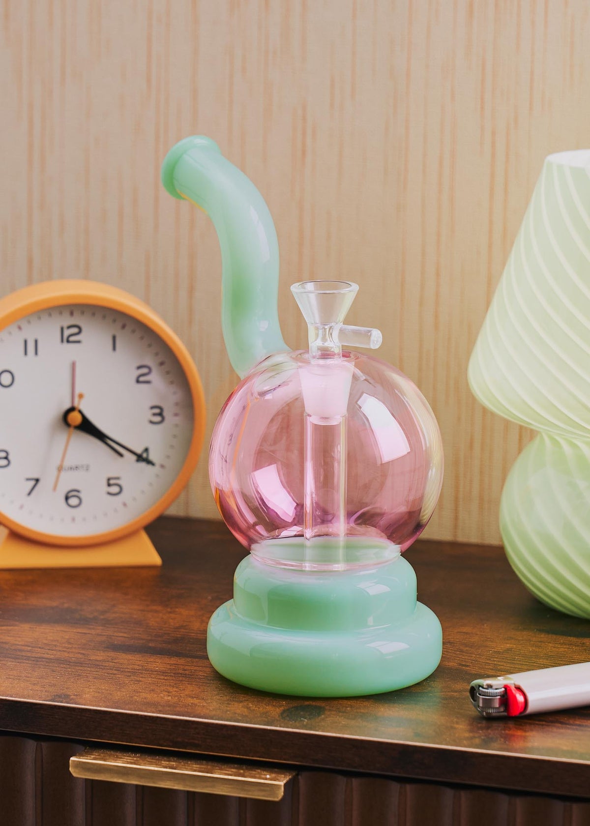 A retro-style squiggle globe bong with a bulbous pink glass body, a light jade green curved neck and base, displayed on a wooden table next to a clock, lighter, and lamp.