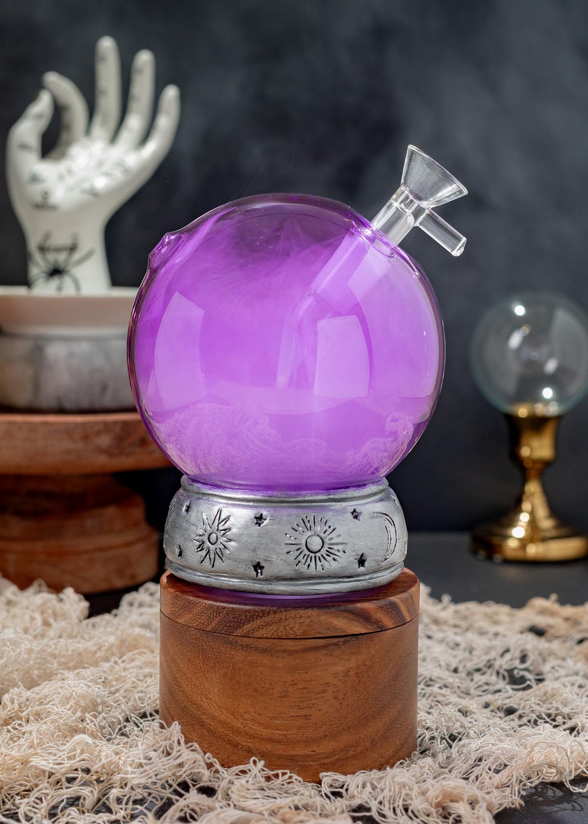 A purple crystal ball bong with a clear glass stopper, resting on a silver celestial-patterned metal band and a wooden base, set against a dark background with mystical props.