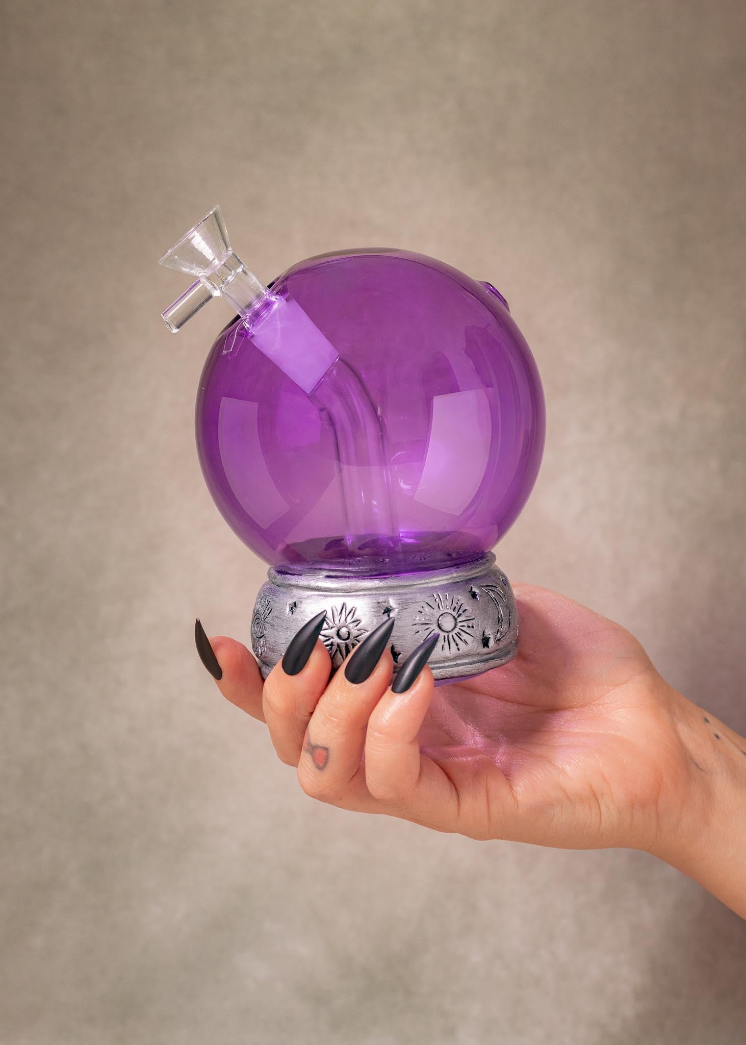 A hand with long black fingernails holds a purple crystal ball glass water pipe with a silver base against a textured beige background.